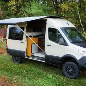 TanVan Campervan auf Mercedes-Sprinter-Basis mit Markise, Outdoor-Küche und robustem Offroad-Setup, geparkt im Grünen.
