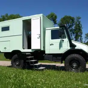 Mintgrünes Expeditionsmobil Exploryx Klippspringer U140L auf Unimog-Basis mit offener Tür, Zugangstreppe und robusten Geländereifen auf einer Wiese