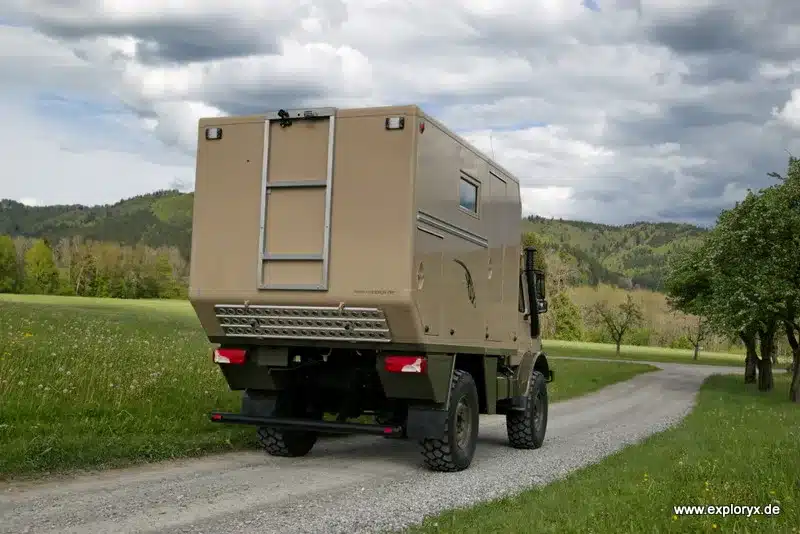 Heckansicht des sandfarbenen Exploryx Klippspringer U1550 L Expeditionsmobils auf einem Feldweg vor bewölktem Himmel und grüner Landschaft.