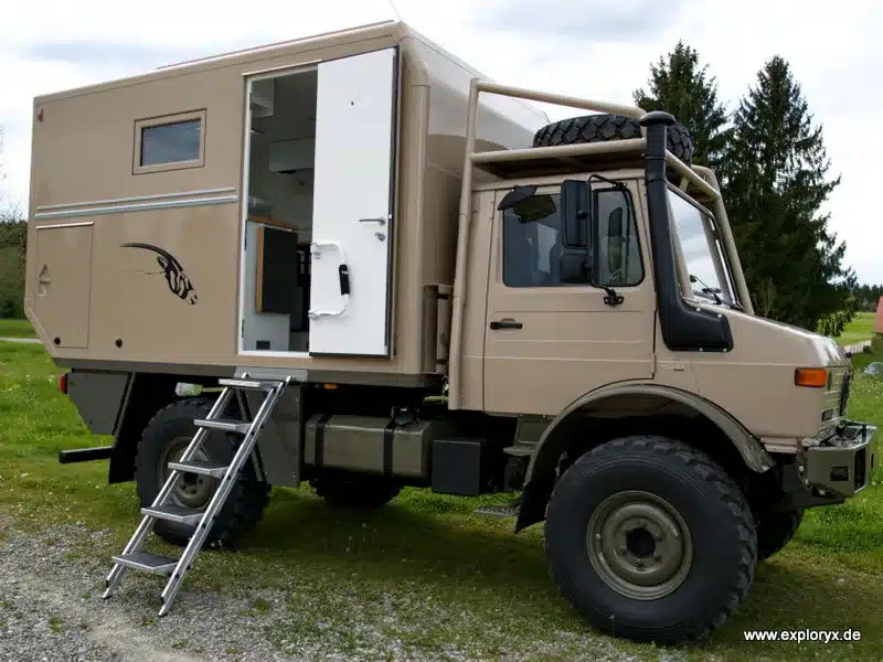 Seitenansicht eines Exploryx Klippspringer U1550 L Expeditionsfahrzeugs auf Unimog-Basis mit geöffneter Einstiegstür und Aluminiumleiter, abgestellt auf einer Wiese.