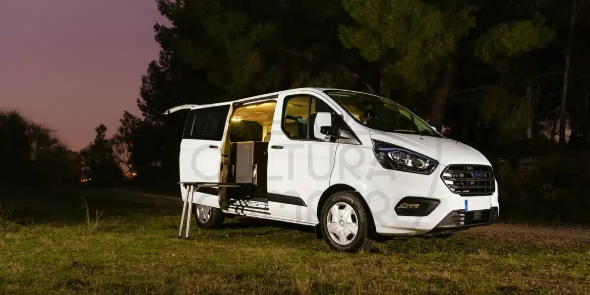 Umgebauter Ford Transit Custom Campervan mit geöffneter Tür und Innenbeleuchtung im Grünen bei Sonnenuntergang.