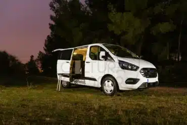 Umgebauter Ford Transit Custom Campervan mit geöffneter Tür und Innenbeleuchtung im Grünen bei Sonnenuntergang.