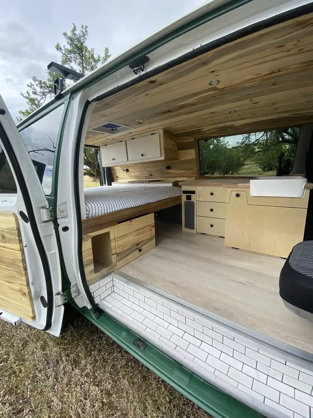 Innenraum eines individuell ausgebauten Vans Green Machine 6AK25 mit Holzverkleidung, Stauraumfächern und festem Bett.