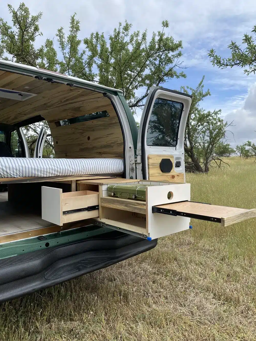 Heckansicht eines Campervans mit ausgezogener Küchenzeile und Stauraum, Green Machine 6AK25, auf einer Wiese.
