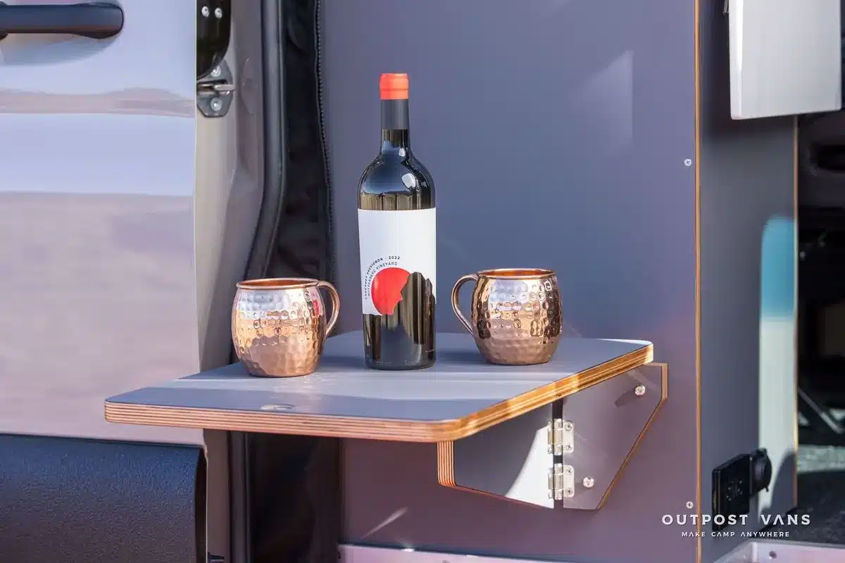 Klappbarer Außentisch am Helix Sprinter 170 AWD Campervan mit zwei Kupferbechern und einer Weinflasche.