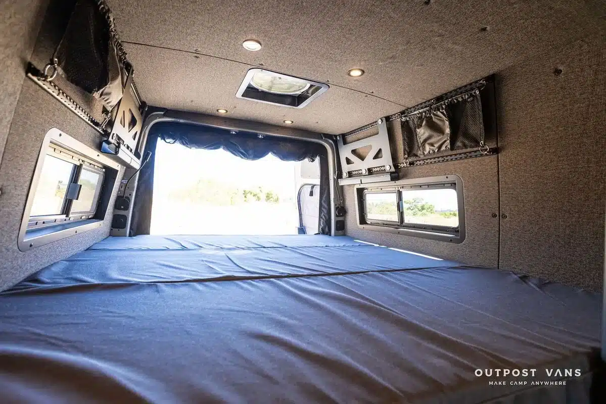 Schlafbereich im Mercedes Sprinter Campervan mit grauer Polsterung, Fenstern und Stauraum.