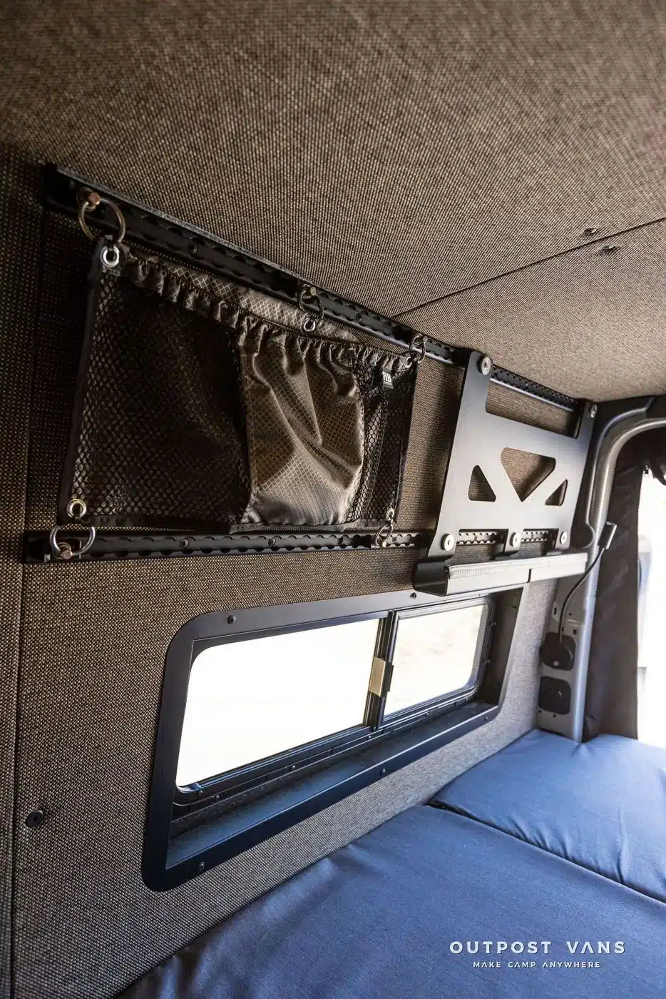 Innenansicht eines Mercedes Sprinter Campervans mit gepolsterten Wandpaneelen, Ablagenetz und Organisationssystem über einem Fenster.