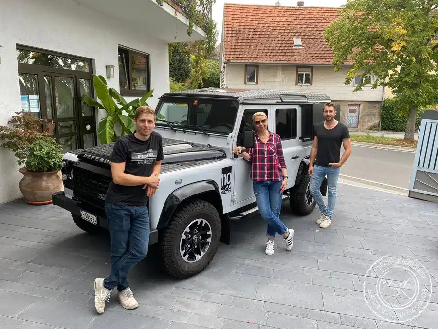Land Rover Defender Camper-Umbau mit drei Personen davor, aufgenommen auf einem Hof in einer Ortschaft.