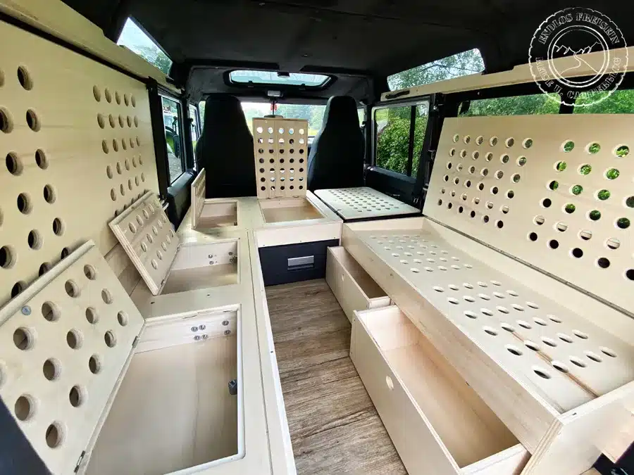 Innenansicht eines zum Camper ausgebauten Land Rover Defender mit aufklappbaren Sitzbänken und integrierten Staufächern aus hellem Holz