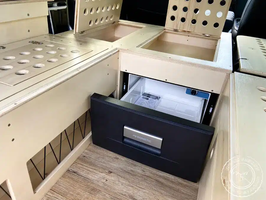 Ausziehbarer Kompressor-Kühlschrank im Holz-Ausbau eines Land Rover Defender Campers mit geöffneten Staufächern.