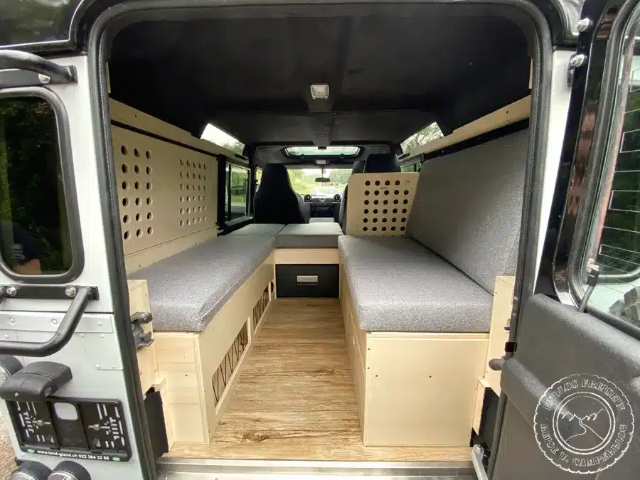 Innenraum eines Land Rover Defender mit maßgefertigtem Holzausbau, zwei Polsterbänken und Stauraum für Campingzwecke.