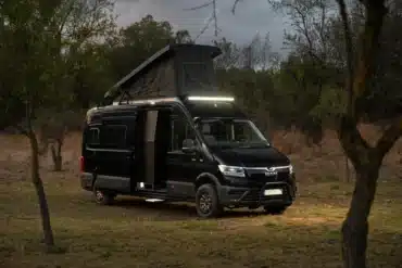 Schwarzer MAN TGE 4x4 Campingbus mit ausgefahrenem Aufstelldach und geöffneter Seitentür, stehend im Gelände am Waldrand.