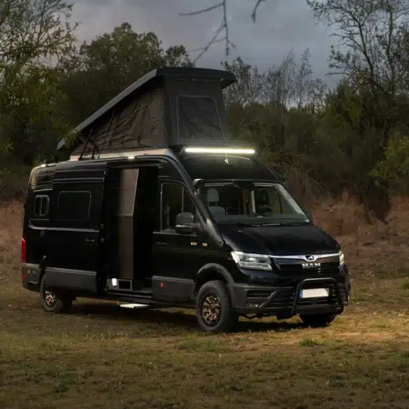 Schwarzer MAN TGE 4x4 Campingbus mit ausgefahrenem Aufstelldach und geöffneter Seitentür, stehend im Gelände am Waldrand.