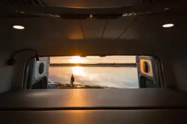 Innenansicht eines Campervans mit Blick auf eine Person am See bei Sonnenuntergang, Türen geöffnet.
