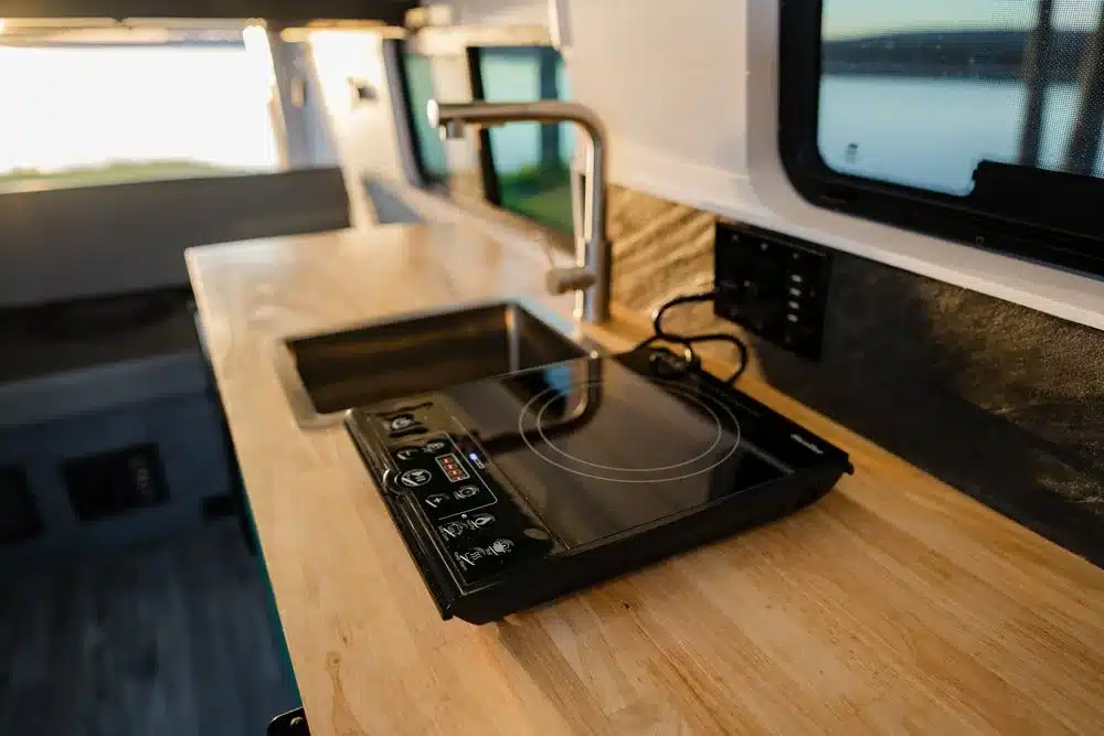 Innenraumansicht einer Campervan-Küchenzeile mit Spüle und Induktionskochfeld, Tageslicht fällt durch das Fenster