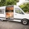 Seitenansicht eines weißen Mercedes Sprinter mit geöffneter Tür, sichtbarem Camper-Ausbau und Sitzgruppe