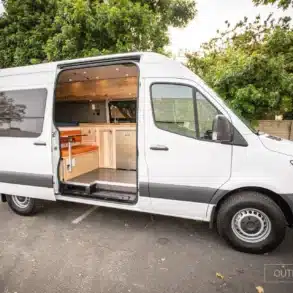 Seitenansicht eines weißen Mercedes Sprinter mit geöffneter Tür, sichtbarem Camper-Ausbau und Sitzgruppe