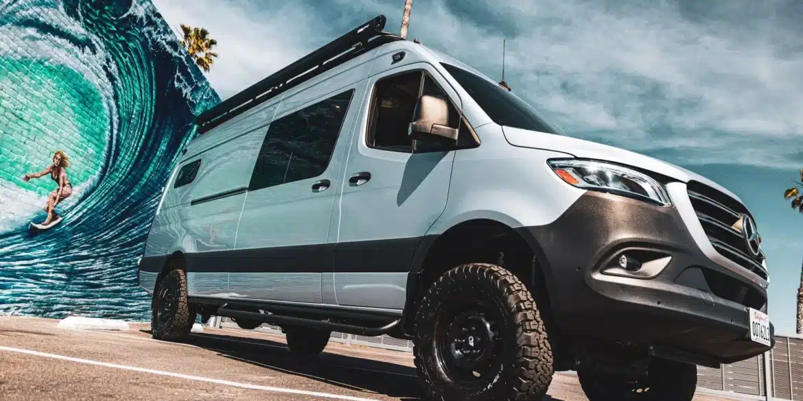 Mercedes Sprinter 4x4 Campervan mit Offroad-Reifen und Dachträger von Outpost Vans vor einer Streetart-Wand mit Surfer.
