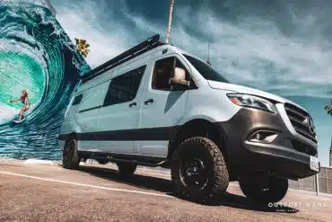 Mercedes Sprinter 4x4 Campervan mit Offroad-Reifen und Dachträger von Outpost Vans vor einer Streetart-Wand mit Surfer.