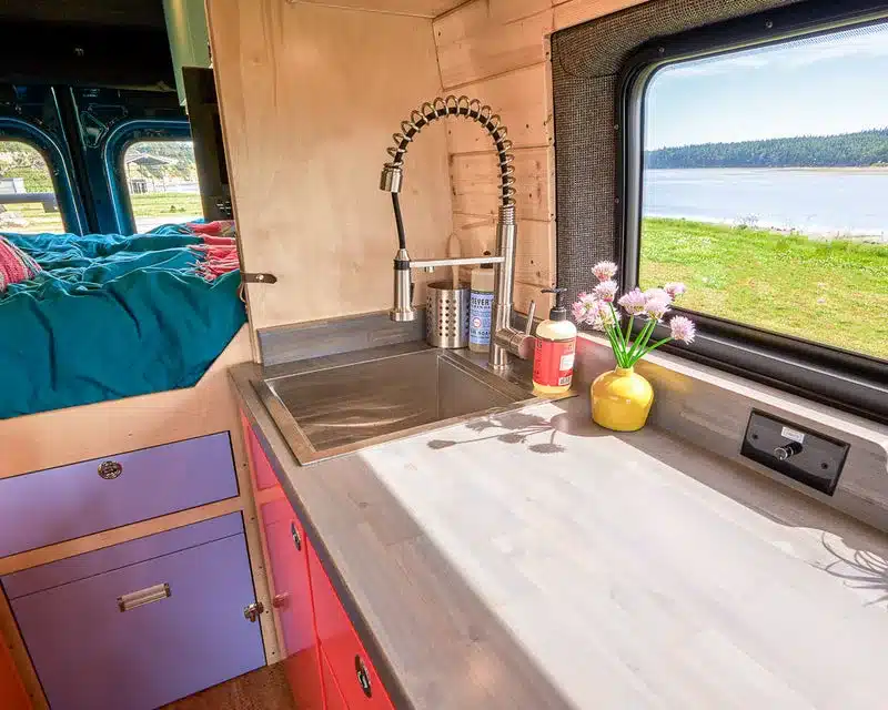 Innenansicht einer Camper-Küche im Mercedes Sprinter mit Spüle, stylischer Armatur, Holzarbeitsplatte und Ausblick auf See und Natur