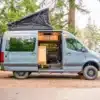 Mercedes Sprinter Family Van mit aufgeklapptem Dachzelt und geöffneten Türen, ausgebaut als Familiencamper, im Grünen geparkt.