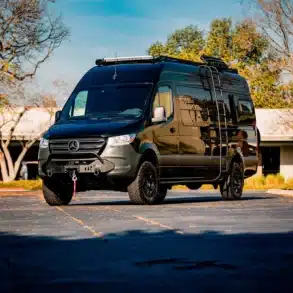 Schwarzer Mercedes Sprinter Mamba Campervan mit Aufbauten und Offroad-Ausstattung auf einem Parkplatz bei sonnigem Wetter