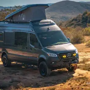 Grauer Mercedes Sprinter Zeus Campervan mit Aufstelldach und Offroad-Umbauten steht auf einem naturbelassenen Feldweg.