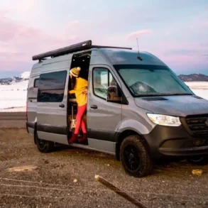 Grauer Mercedes Sprinter Camper-Van mit Markise, geöffnetem Seiteneingang und Person, stehend in der Schiebetür; im Hintergrund schneebedeckte Berge und offene Landschaft.