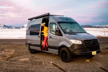 Grauer Mercedes Sprinter Camper-Van mit Markise, geöffnetem Seiteneingang und Person, stehend in der Schiebetür; im Hintergrund schneebedeckte Berge und offene Landschaft.