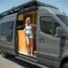 Frau steht in geöffneter Tür eines hochwertig ausgebauten, grauen Camper-Vans Moose mit sichtbaren Holzelementen und Dachterrasse