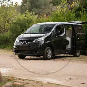 Schwarzer Nissan NV200 als Camper mit geöffneter Schiebetür auf einem Naturparkplatz.