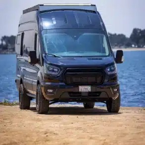 ONYX Transit Campervan auf Ford-Transit-Basis, mit Dachträger und Offroad-Bereifung, am Ufer eines Sees geparkt.