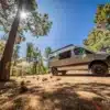 Outpost Vans CRAG Offroad-Campervan auf Basis eines Mercedes-Benz Sprinter 144 AWD, geparkt im Wald bei Sonnenschein, bereit für Abenteuer abseits befestigter Wege.