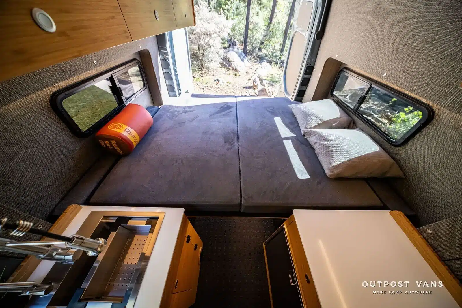 Schlafbereich mit festem Bett, Kissen und seitlichen Fenstern im Outpost Vans CRAG Campervan, vorne Küchenelemente aus Bambus und Corian