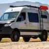 DUNE Campervan, ausgebauter Mercedes Sprinter 4x4 Offroad mit Dachträger, Leiter und Expeditionszubehör auf Schotterpiste.