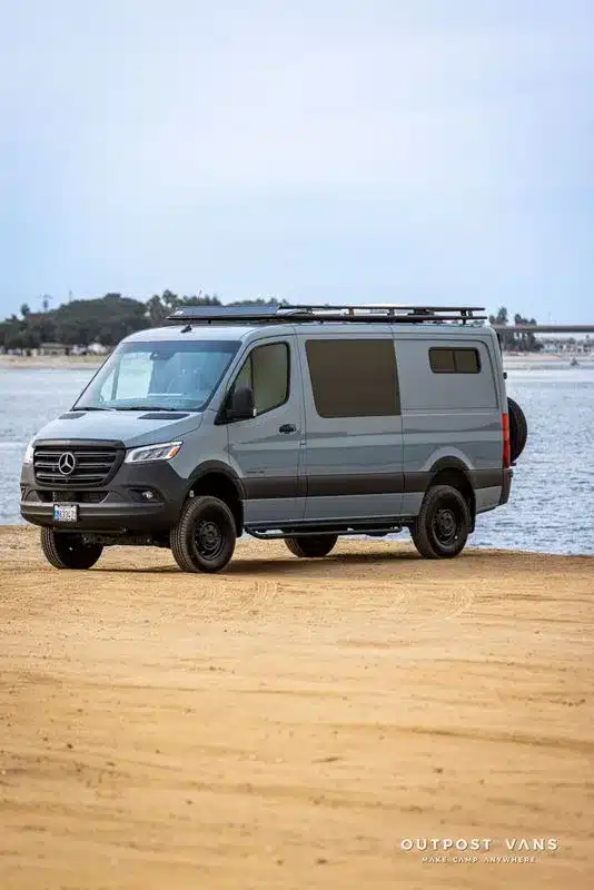 Outpost Vans Jasper Campervan auf Mercedes Sprinter 144 mit Dachträger, niedrigen Dach und Reserverad, am Sandstrand geparkt.