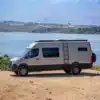 Seitenansicht eines silbernen Mercedes Sprinter 170 High Roof Campervans mit Dachträger, Leiter und Markise am Wasser.