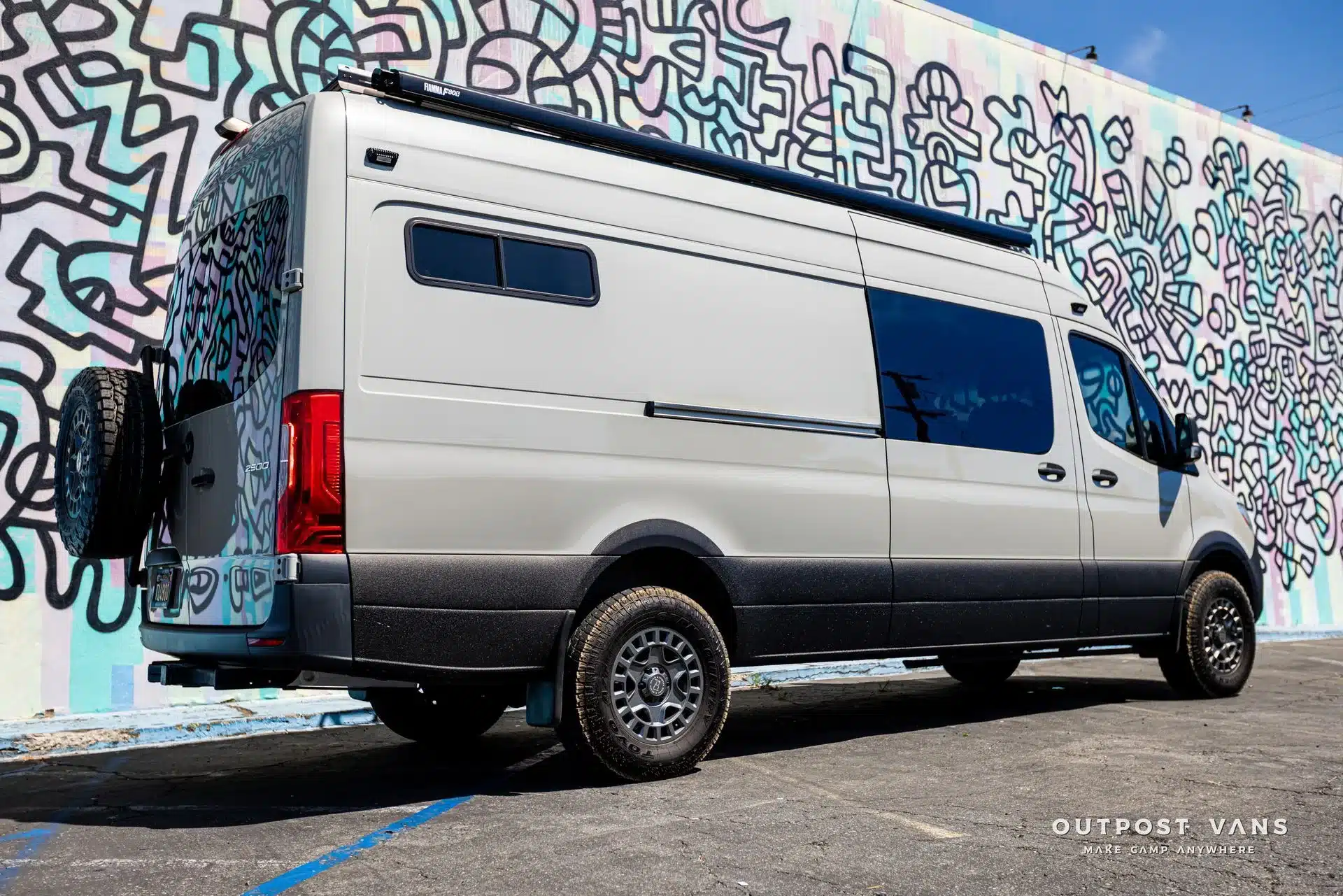 Mercedes Sprinter 170 High Roof Campervan Jerry von Outpost Vans mit Markise und Geländereifen vor einer bunten Graffiti-Wand.