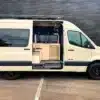 Outpost Vans OPV-04 Campervan mit offener Tür, sichtbarem Innenausbau und moderner Ausstattung, stehend auf einem Parkplatz.