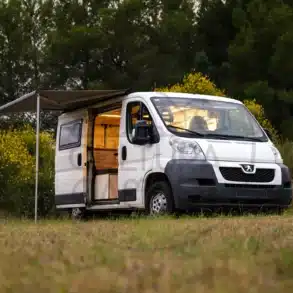 Peugeot Boxer L1H1 Van-Cabaña Camper mit Markise, geöffnetem Seiteneingang und beleuchtetem Innenraum auf einer Wiese vor Bäumen.