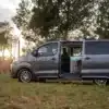 Peugeot Traveller Campervan mit geöffnetem Seiteneinstieg, parkend auf einer Wiese in der Natur, Innenausbau sichtbar.
