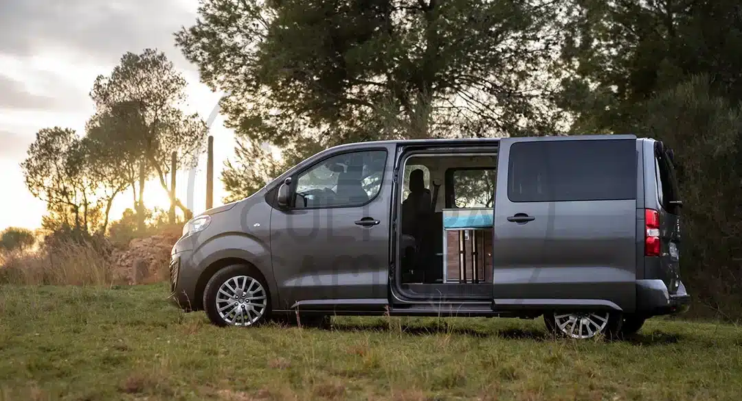 Peugeot Traveller Campervan mit geöffnetem Seiteneinstieg, parkend auf einer Wiese in der Natur, Innenausbau sichtbar.