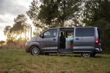 Peugeot Traveller Campervan mit geöffnetem Seiteneinstieg, parkend auf einer Wiese in der Natur, Innenausbau sichtbar.