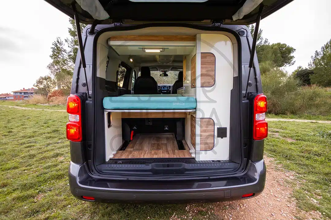 Innenansicht eines Peugeot Traveller Campers mit Bett und Stauraum, aufgenommen von der geöffneten Heckklappe aus.