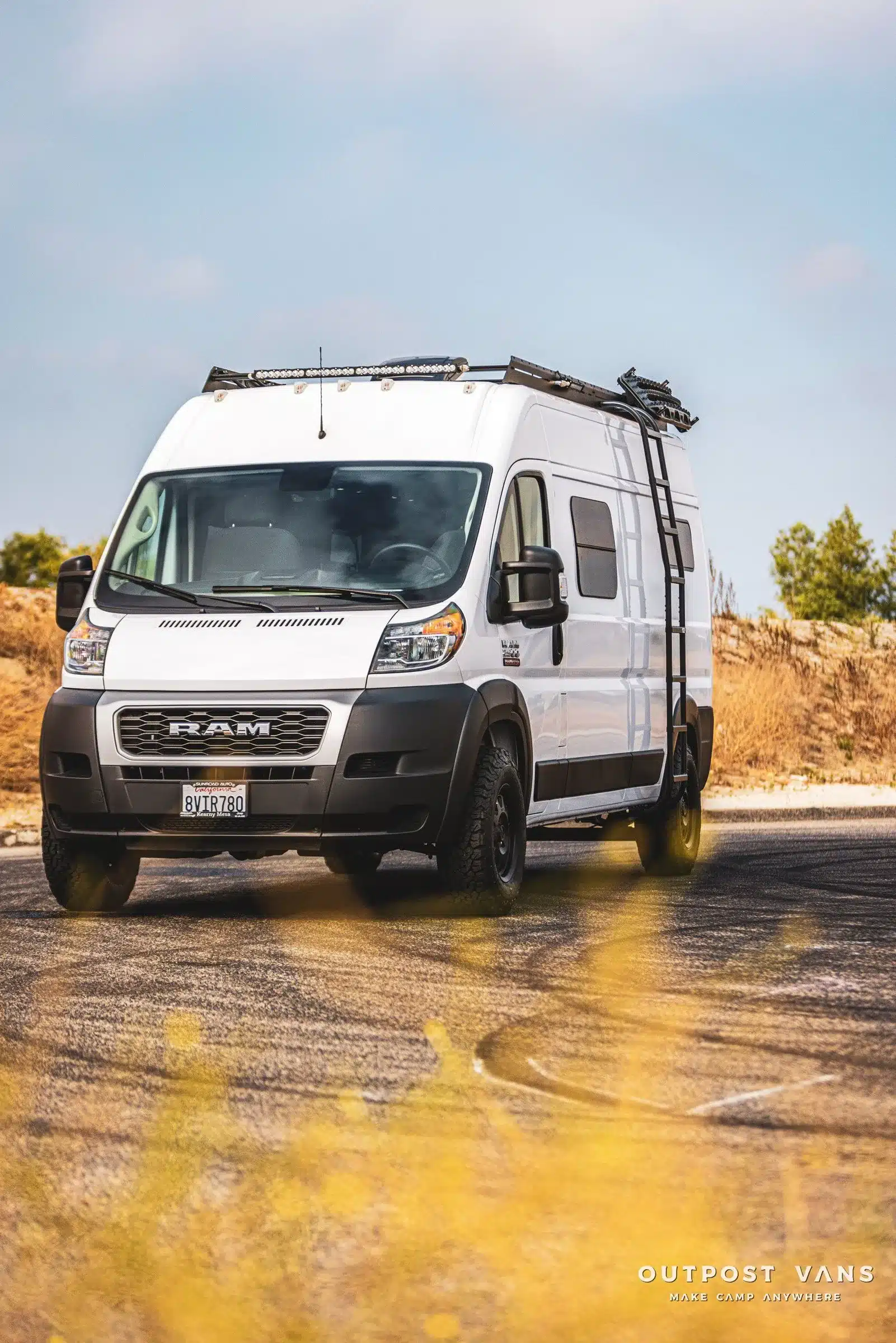 Weißer Ram Promaster Altitude Campervan mit Dachträger, Leiter und Offroad-Ausstattung auf einer Straße vor natürlicher Kulisse.