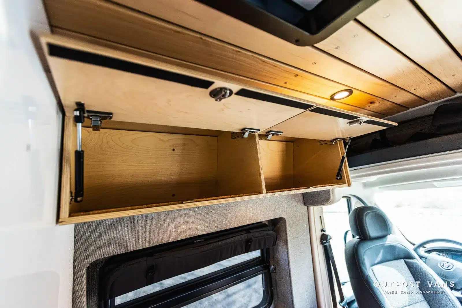 Offener, leerer Oberschrank aus Holz mit Fächern im Wohnbereich eines modernen US-Campervans, darunter ein Fenster und Fahrersitz sichtbar.
