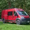 Roter Ram ProMaster Campervan steht auf einer grünen Wiese vor einem Wald.