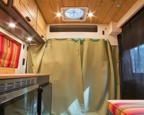 Innenraum eines ausgebauten Ram ProMaster mit Küchenzeile, Holzboden, Oberschränken und Vorhang.