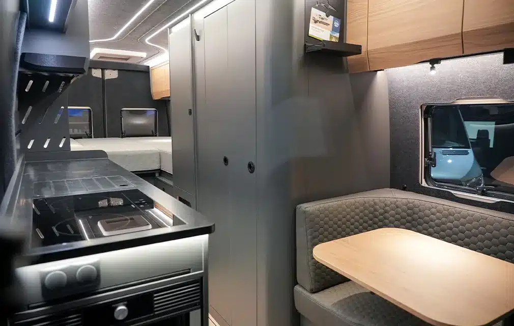 Innenansicht eines ausgebauten Mercedes Sprinter Campervans mit Küchenzeile, Sitzgruppe und Schlafbereich.
