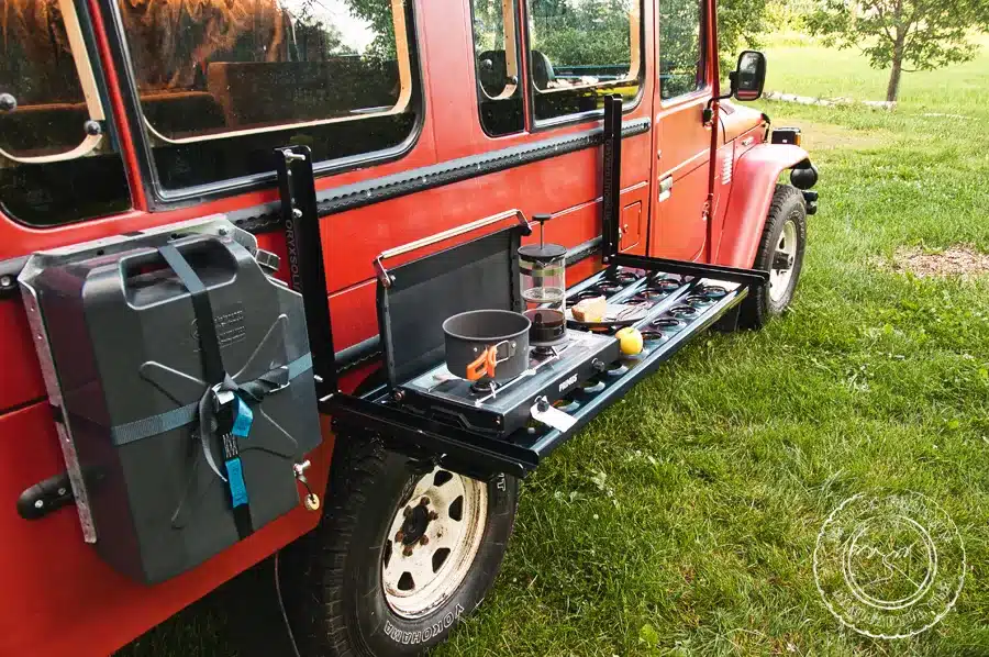 Klappbare Außenküche mit Kanistern und Gaskocher an einem roten Toyota Land Cruiser BJ45 auf einer Wiese.